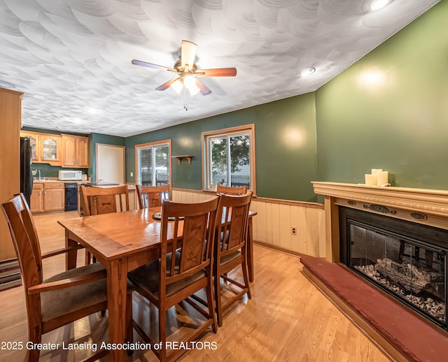 dining area featuring ceiling fan, a textured ceiling, a wainscoted wall, light wood-type flooring, and a glass covered fireplace