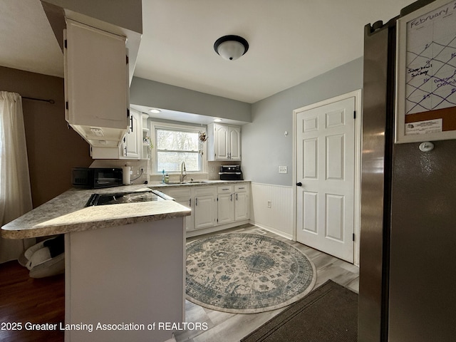 kitchen featuring white cabinets, wainscoting, a peninsula, black microwave, and a sink