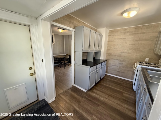 kitchen with dark wood-style flooring, dark countertops, brick wall, baseboards, and white gas range oven