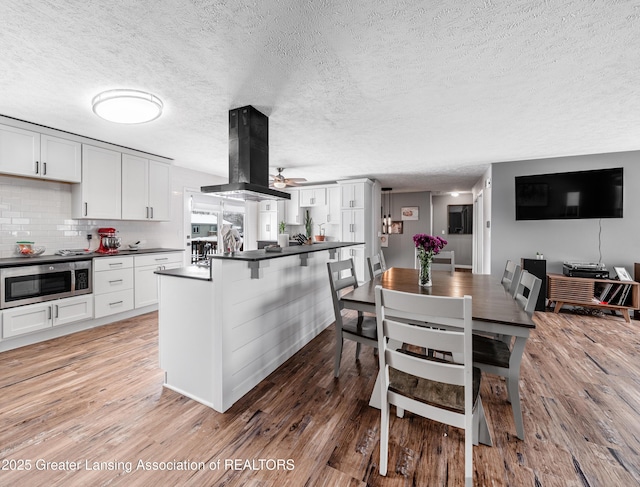 kitchen with tasteful backsplash, dark countertops, stainless steel microwave, light wood-style flooring, and island range hood