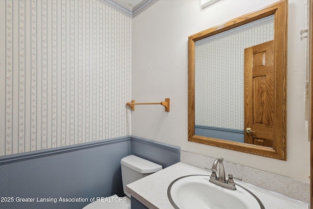 half bathroom featuring toilet, wallpapered walls, vanity, and wainscoting