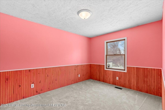 carpeted empty room with a wainscoted wall, a textured ceiling, visible vents, and wooden walls