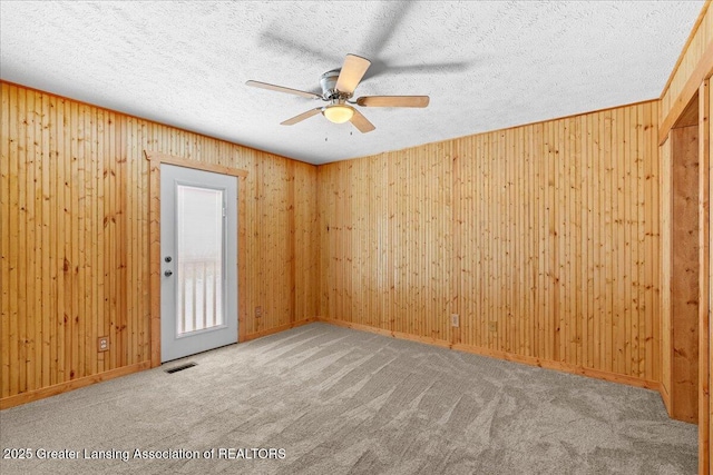 unfurnished room with a textured ceiling, ceiling fan, carpet floors, wood walls, and visible vents