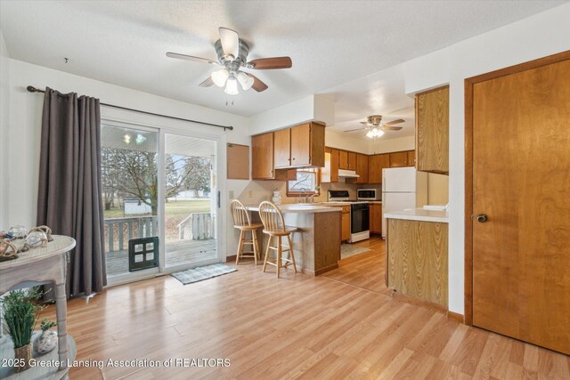 kitchen featuring under cabinet range hood, a peninsula, white appliances, light countertops, and brown cabinets