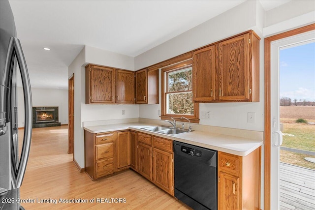 kitchen featuring a warm lit fireplace, light wood-style flooring, a sink, refrigerator with ice dispenser, and black dishwasher