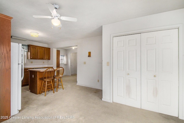 kitchen with a breakfast bar area, light colored carpet, light countertops, freestanding refrigerator, and brown cabinets