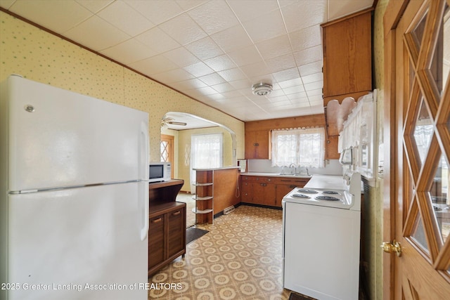 kitchen with arched walkways, white appliances, a sink, light floors, and wallpapered walls