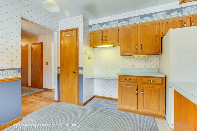 kitchen featuring wallpapered walls, baseboards, brown cabinets, freestanding refrigerator, and light countertops