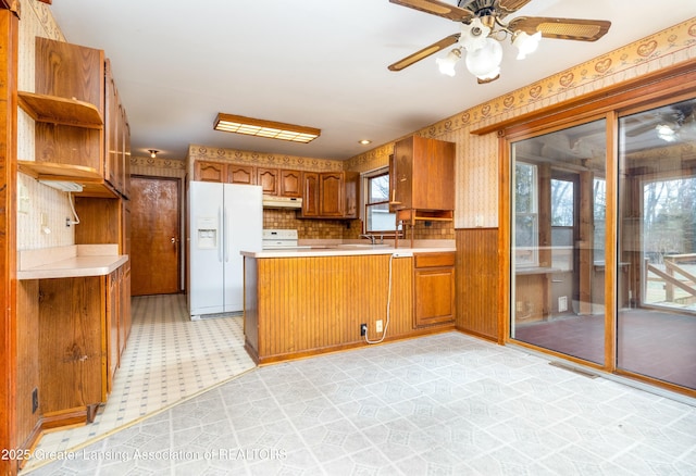 kitchen with wallpapered walls, light floors, a peninsula, white refrigerator with ice dispenser, and brown cabinetry