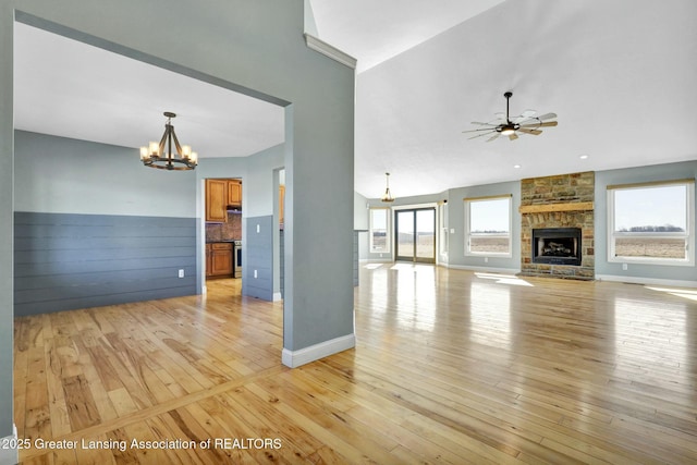 unfurnished living room with light wood finished floors, a stone fireplace, ceiling fan with notable chandelier, and baseboards