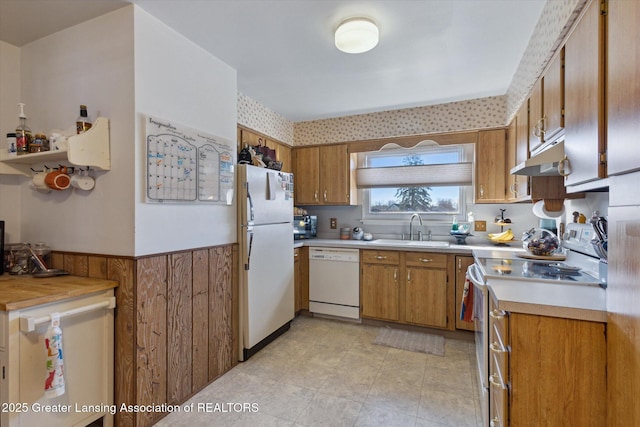 kitchen with under cabinet range hood, a wainscoted wall, brown cabinets, white appliances, and a sink