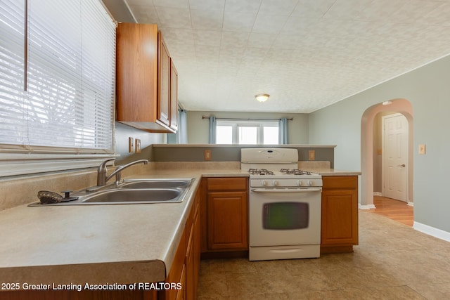 kitchen featuring a peninsula, arched walkways, a sink, white gas range oven, and brown cabinets