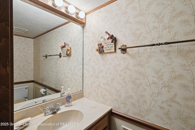 half bathroom featuring a textured ceiling, toilet, vanity, and wallpapered walls