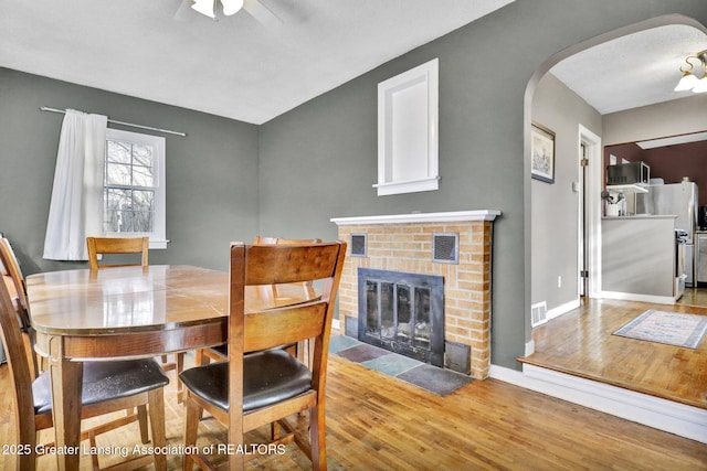 dining area featuring a ceiling fan, wood finished floors, baseboards, arched walkways, and a brick fireplace