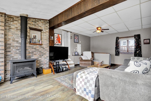 living room featuring a ceiling fan, a drop ceiling, wood finished floors, baseboards, and a wood stove