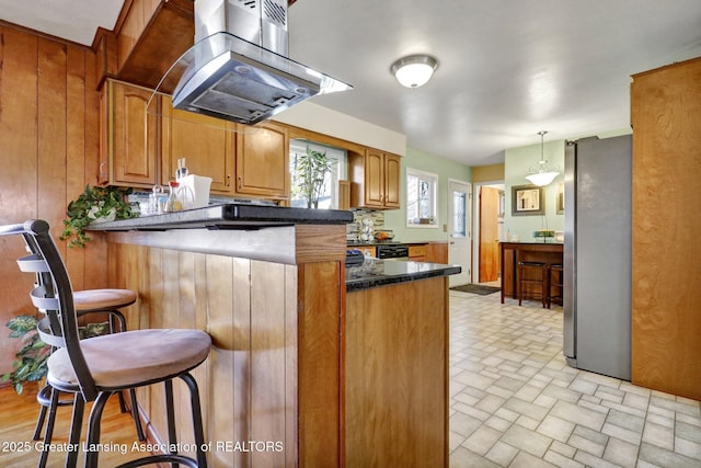 kitchen featuring a kitchen bar, brown cabinets, freestanding refrigerator, wooden walls, and island range hood