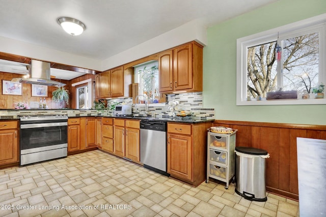 kitchen featuring a wainscoted wall, stone finish floor, island exhaust hood, stainless steel appliances, and brown cabinetry