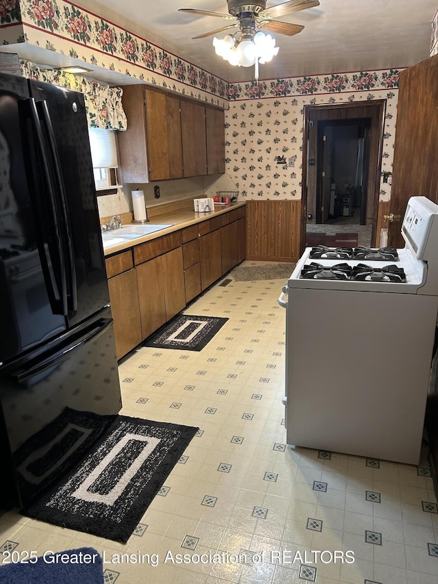 kitchen featuring wallpapered walls, gas range gas stove, a wainscoted wall, freestanding refrigerator, and a ceiling fan