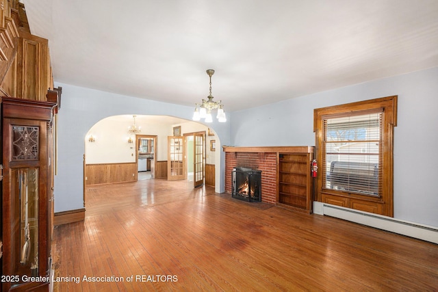 unfurnished living room featuring arched walkways, wainscoting, a fireplace, a baseboard radiator, and a chandelier