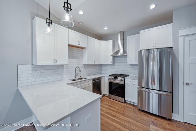 kitchen with wall chimney range hood, appliances with stainless steel finishes, wood finished floors, white cabinetry, and a sink