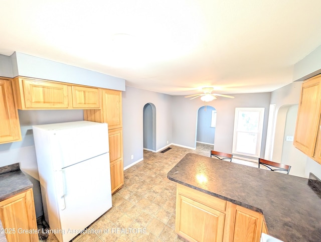 kitchen with freestanding refrigerator, a peninsula, light brown cabinets, dark countertops, and arched walkways