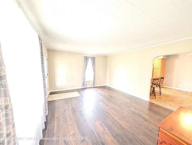 empty room featuring arched walkways, crown molding, baseboards, and wood finished floors