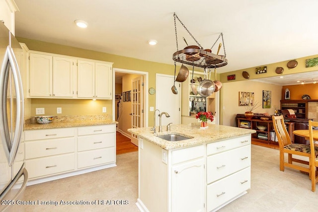 kitchen featuring freestanding refrigerator, light stone countertops, a sink, white cabinets, and a kitchen island with sink
