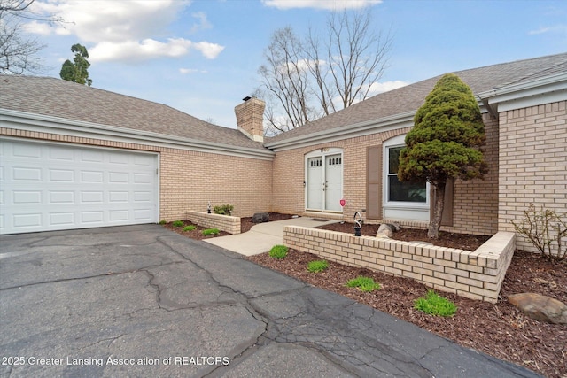 view of front facade with a chimney, aphalt driveway, a shingled roof, an attached garage, and brick siding