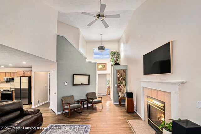 living area with ceiling fan, baseboards, a tile fireplace, light wood-type flooring, and a towering ceiling