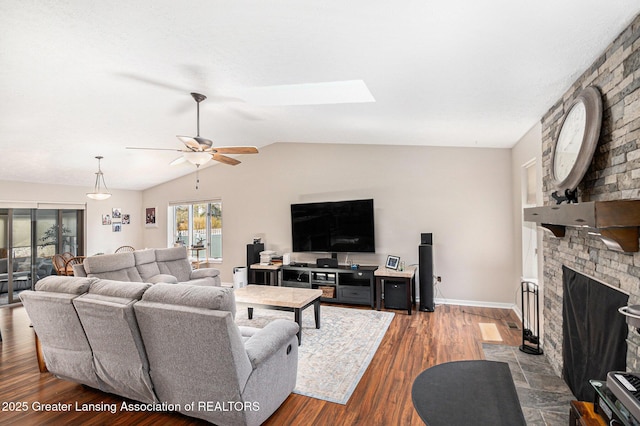 living area with vaulted ceiling with skylight, a brick fireplace, ceiling fan, baseboards, and dark wood finished floors