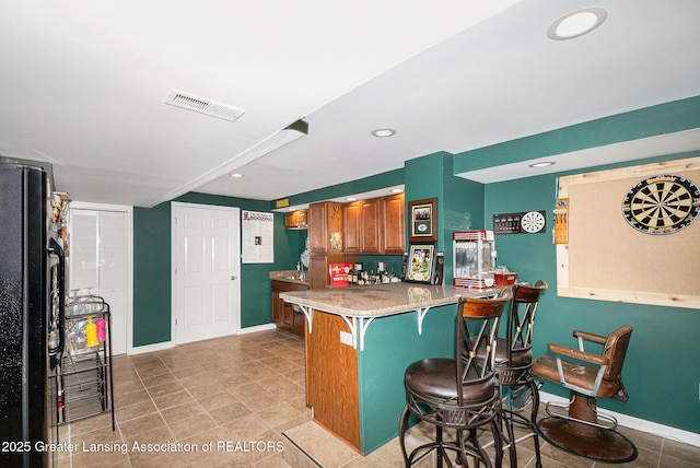 kitchen featuring freestanding refrigerator, brown cabinetry, a kitchen bar, baseboards, and visible vents