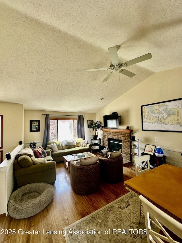 living area featuring lofted ceiling, a textured ceiling, ceiling fan, a brick fireplace, and wood finished floors