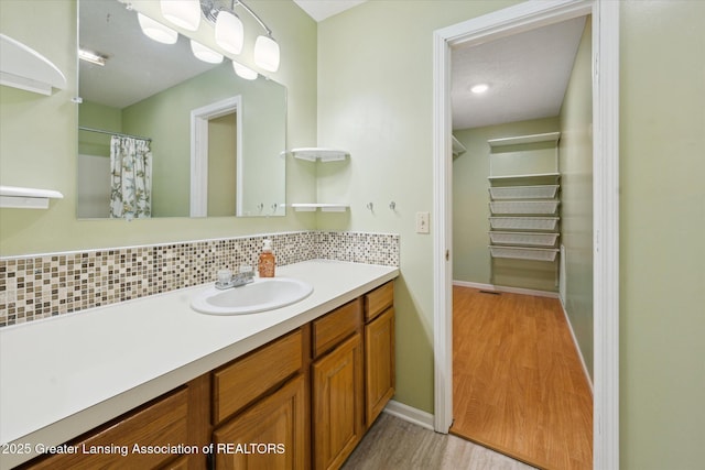 full bath with a walk in closet, vanity, baseboards, wood finished floors, and backsplash