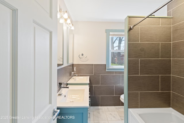 bathroom featuring double vanity, tile walls, a sink, a wainscoted wall, and toilet