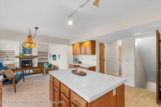 kitchen featuring brown cabinets, tile countertops, a warm lit fireplace, and light wood-style flooring