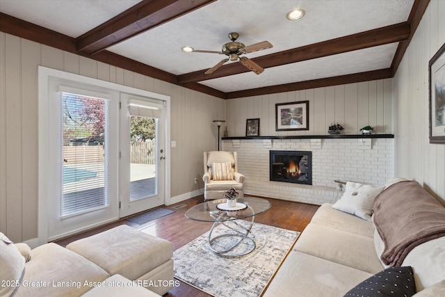 living room featuring ceiling fan, a brick fireplace, beam ceiling, and wood finished floors