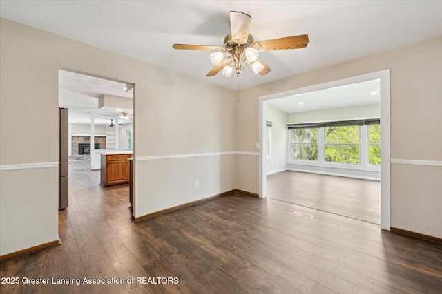 unfurnished room featuring dark wood-style flooring, ceiling fan, a fireplace with raised hearth, baseboards, and visible vents
