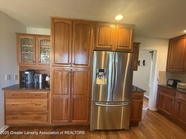 kitchen featuring brown cabinetry, wood finished floors, glass insert cabinets, stainless steel fridge, and dark stone counters