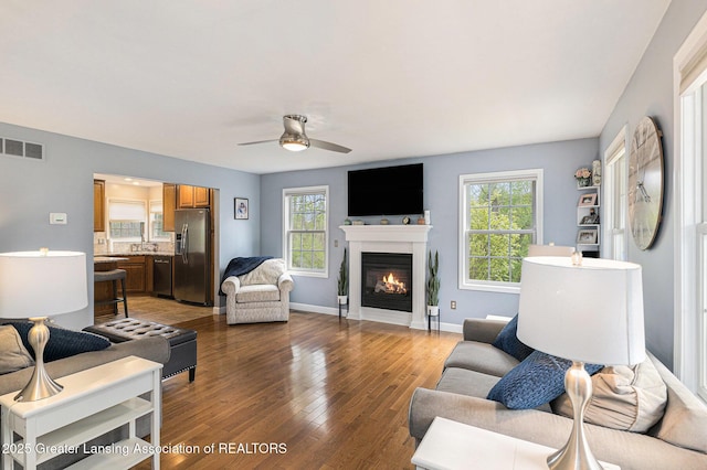 living area featuring a ceiling fan, visible vents, baseboards, light wood finished floors, and a glass covered fireplace