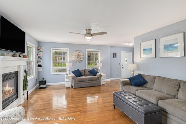 living area featuring a glass covered fireplace, baseboards, a ceiling fan, and light wood-type flooring