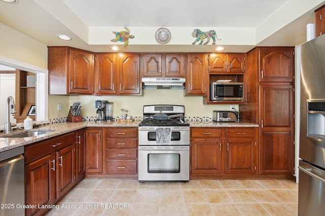 kitchen featuring light stone counters, a sink, brown cabinetry, stainless steel appliances, and under cabinet range hood