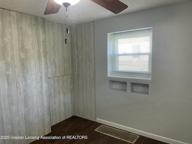 empty room featuring a textured ceiling, a ceiling fan, baseboards, visible vents, and dark wood-style floors