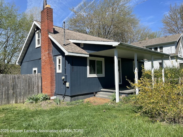 back of house with covered porch, a chimney, fence, a yard, and a shingled roof