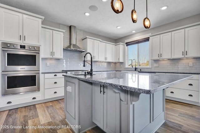 kitchen with wall chimney range hood, backsplash, a center island with sink, a sink, and double oven