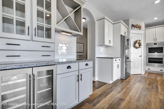 kitchen with arched walkways, stone countertops, stainless steel appliances, wine cooler, and dark wood-style floors
