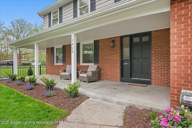 view of exterior entry featuring covered porch, brick siding, and fence