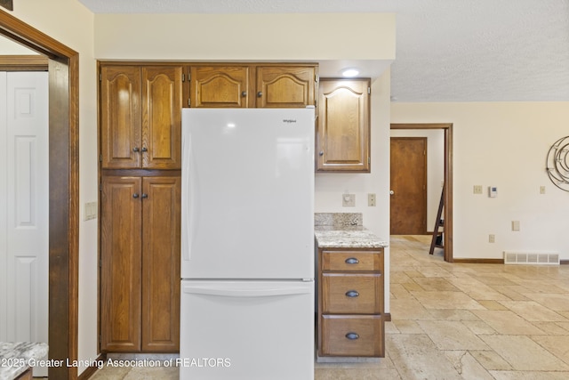 kitchen with freestanding refrigerator, baseboards, stone tile floors, brown cabinets, and light stone counters