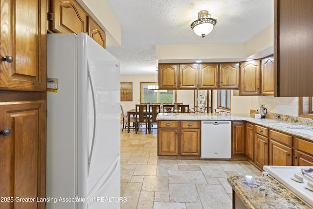 kitchen featuring white appliances, brown cabinetry, a textured ceiling, stone tile floors, and a peninsula