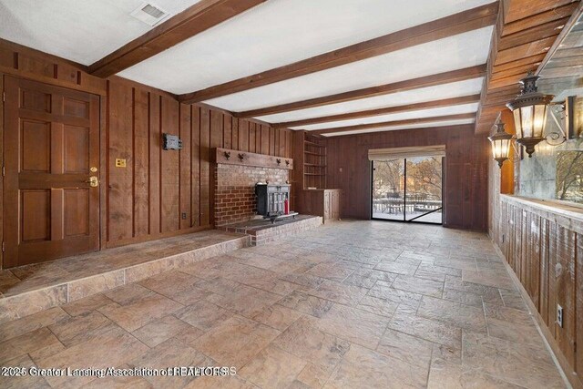 unfurnished living room with wooden walls, stone tile floors, beamed ceiling, and a wood stove