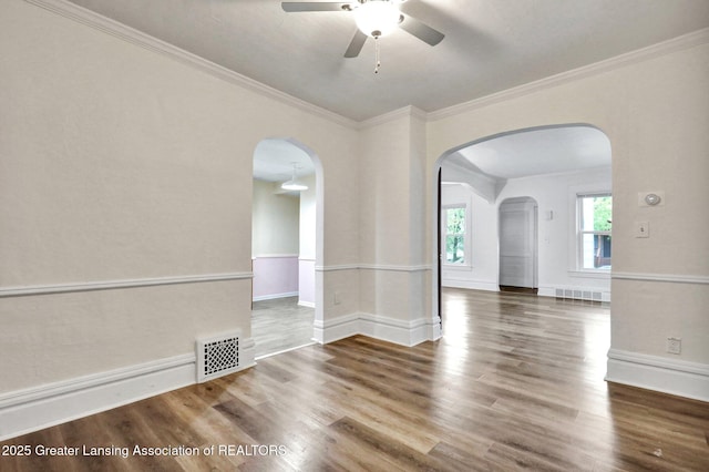 empty room with arched walkways, a ceiling fan, wood finished floors, crown molding, and baseboards